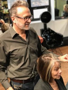 A hairstylist with glasses expertly applies vibrant hair color as she stands beside a seated woman in the salon, carefully styling her hair to complement her skin tone. - Pompadours Hair Salon | Boulder, CO