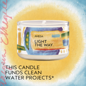 A glass candle labeled "Aveda Light the Way" with colorful artwork; text below states, "This candle funds clean water projects. - Pompadours Hair Salon | Boulder, CO