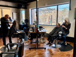 Two hairstylists work on clients’ hair in a modern salon with wooden floors, large windows, and various hair tools on a central station. - Pompadours Hair Salon | Boulder, CO